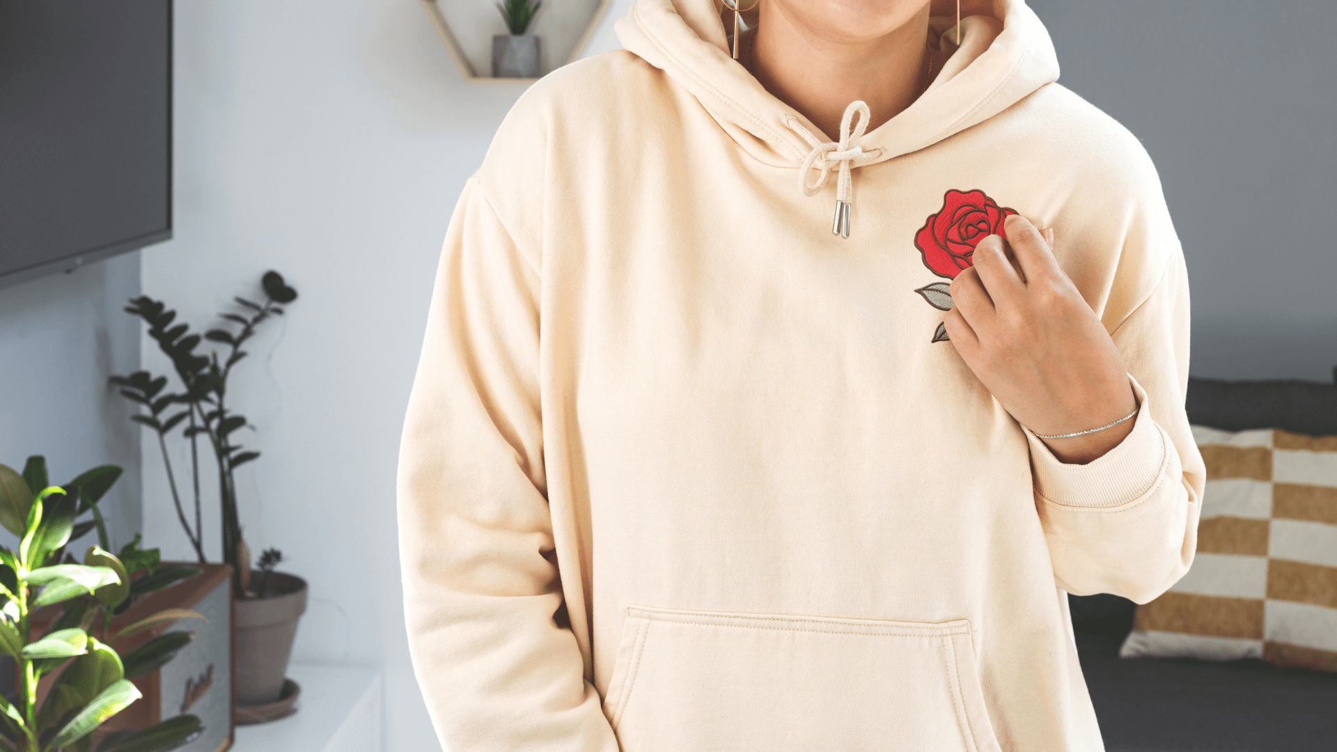 a person wearing a light hoodie with an embroidered rose over the left chest that they are touching with their hand