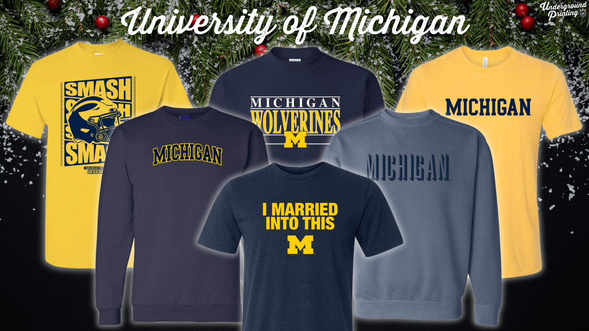 A collection of University of Michigan apparel against a holiday-themed backdrop, including t-shirts and sweatshirts in classic navy and maize colors with "Michigan Wolverines," "I Married Into This," and other Michigan-themed designs.