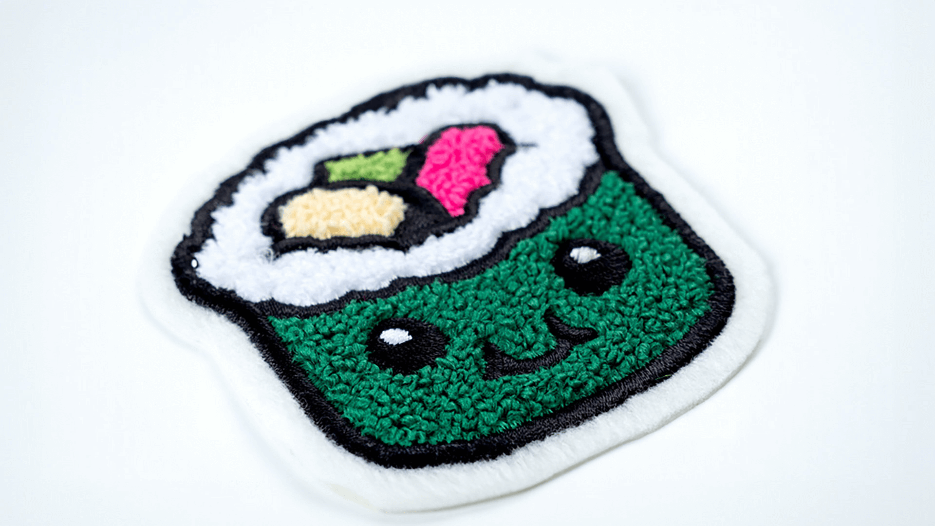 A cute chenille patch designed as a smiling sushi roll with a green exterior, white rice border, and colorful filling. The patch has a soft, fuzzy texture with black outlining on a white background.