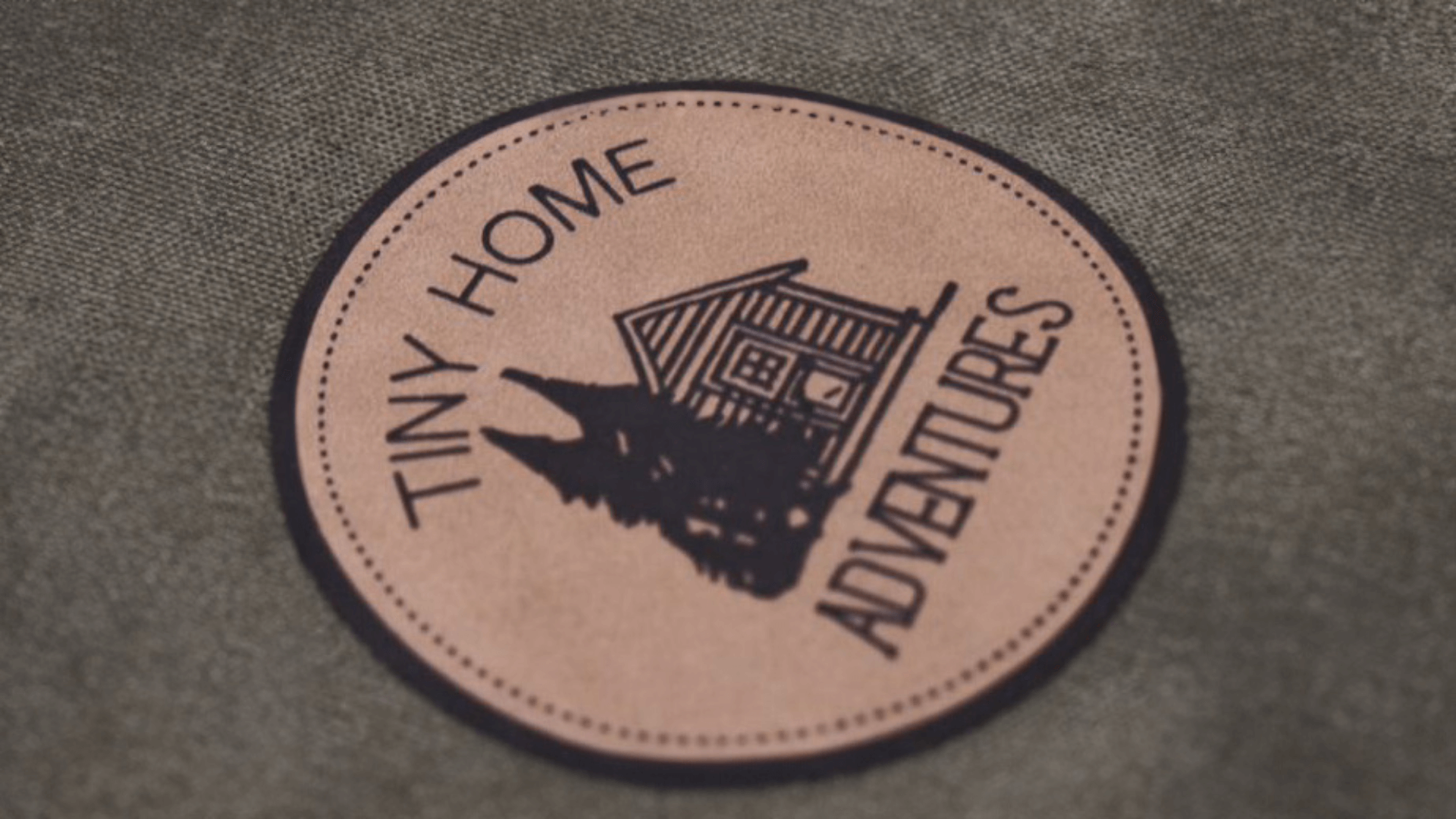 A circular faux leather patch with "Tiny Home Adventures" written around an illustration of a small cabin surrounded by trees. The design is printed in black on a tan faux leather background with stitched edges