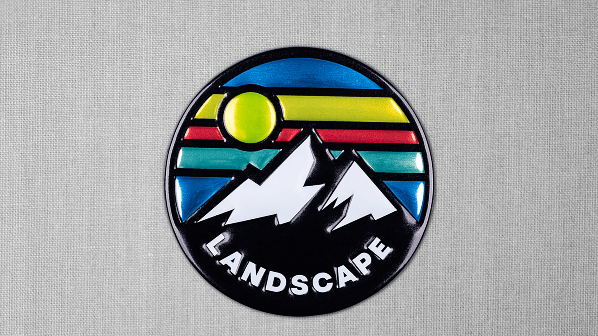 A glossy, circular patch depicting a stylized landscape with two white mountain peaks, a yellow sun, and colorful stripes in green, red, and blue, with the word "LANDSCAPE" at the bottom, set against a gray fabric background