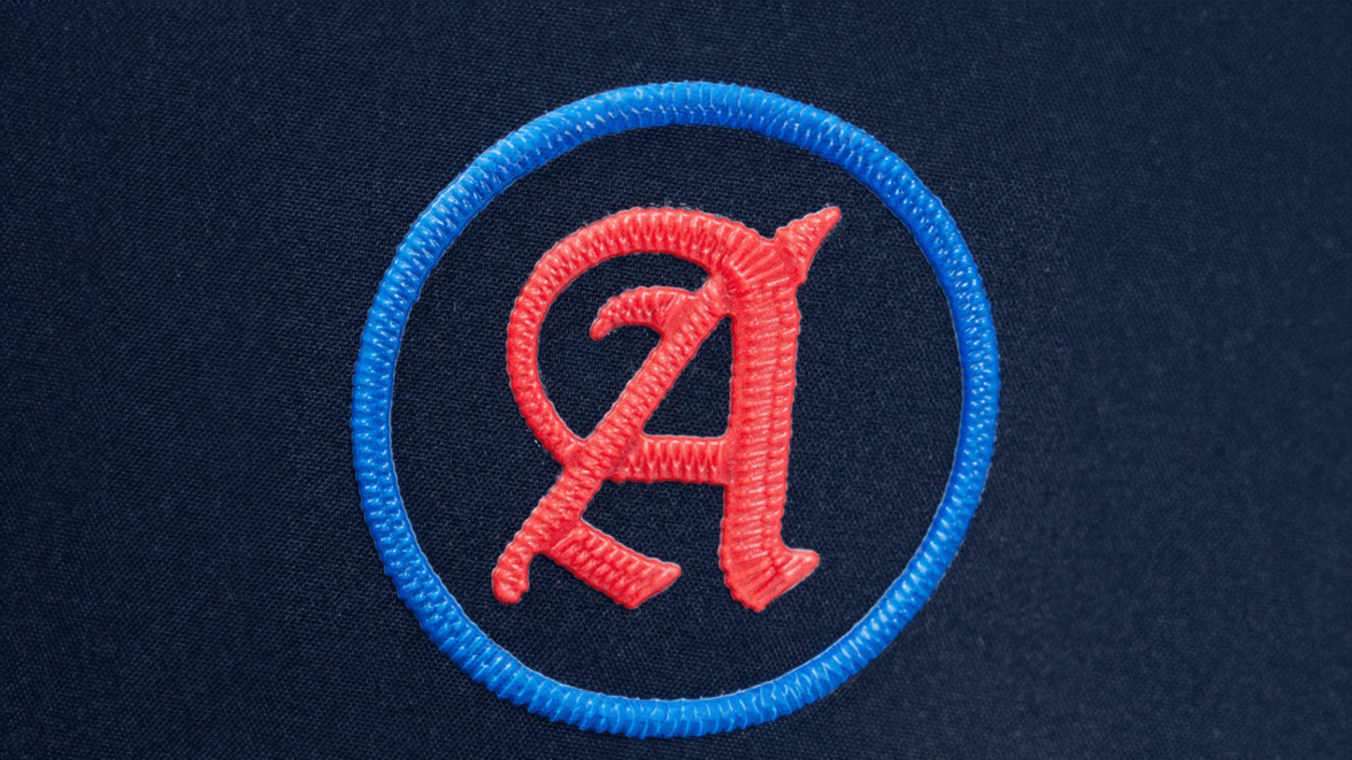 A simple, circular patch with a red letter "A" inside a blue ring, sewn onto a dark navy blue fabric background