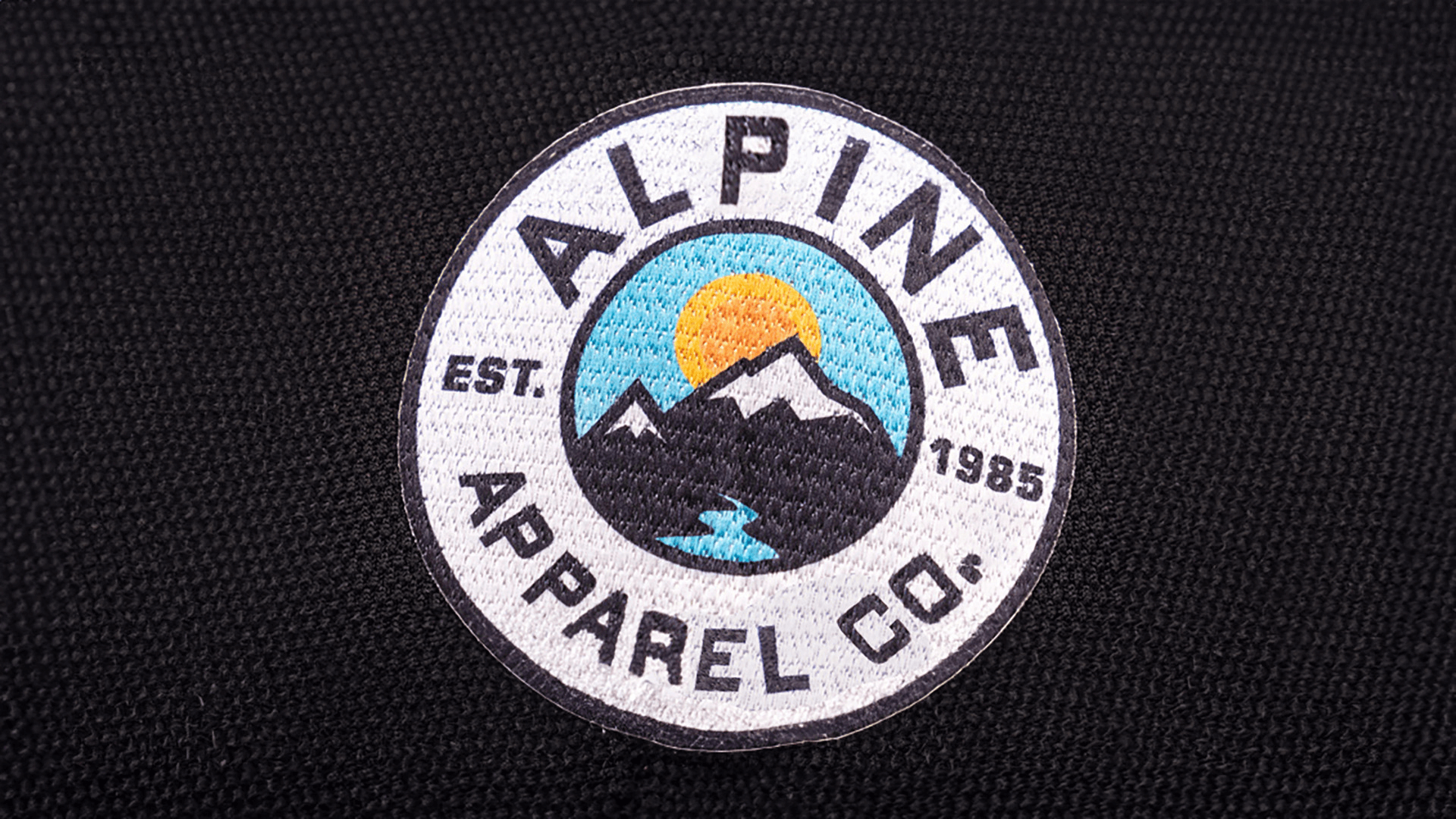 A circular patch for "Alpine Apparel Co." with an illustrated mountain scene, featuring a rising sun and a river in blue and yellow tones, framed by the text "Alpine Apparel Co. Est. 1985" on a black fabric background