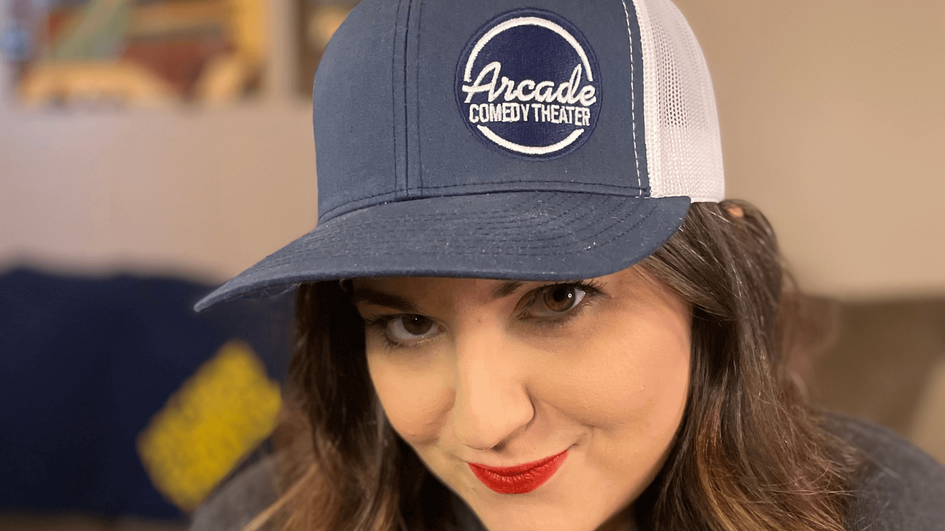 A girl smiling wearing a custom trucker hat with an embroidered patch for the arcade comedy theatre brand 