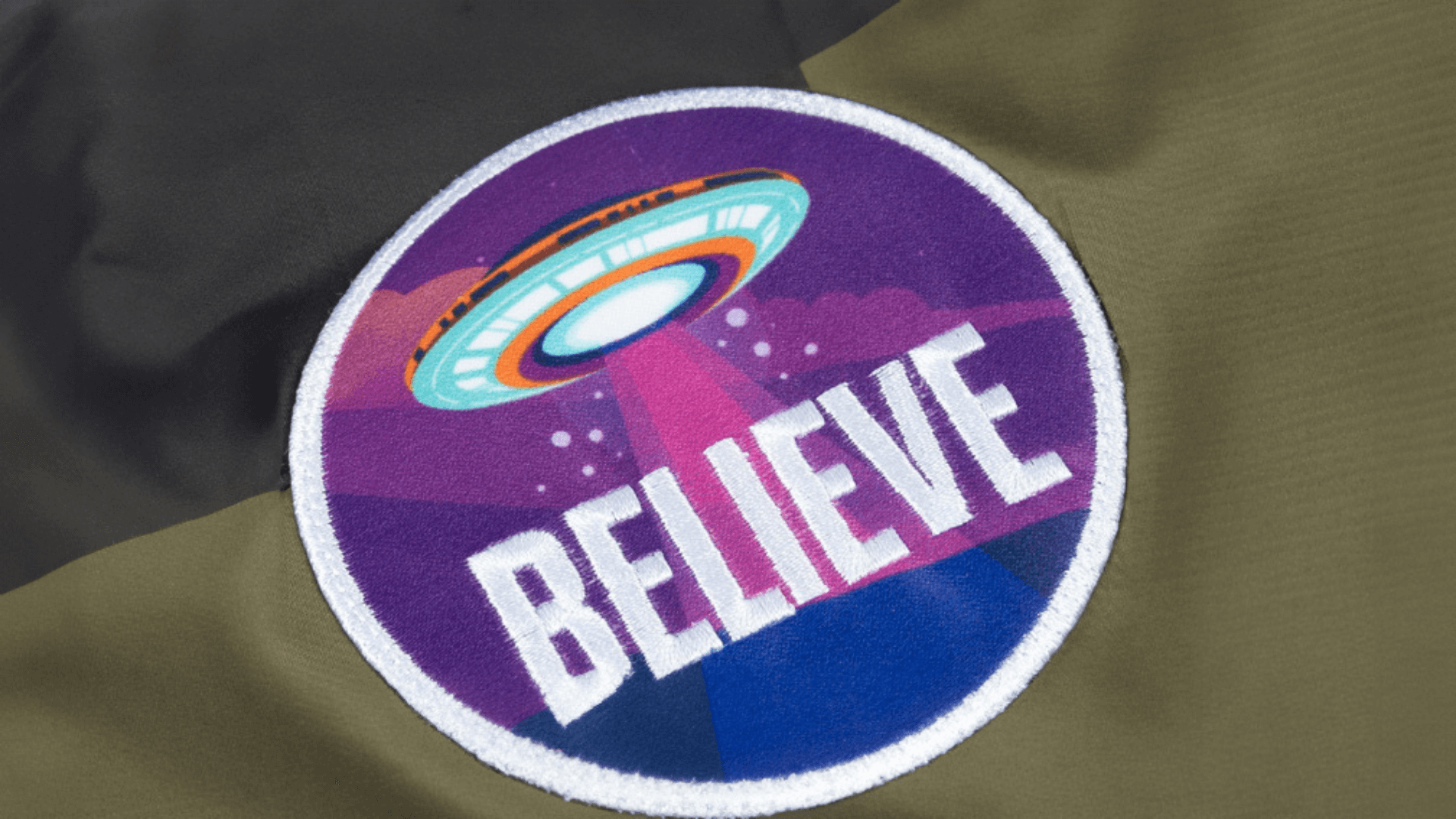  A colorful circular patch with a UFO beaming light downwards, with the word "BELIEVE" in bold white letters, set against a purple background and sewn onto an olive green jacket