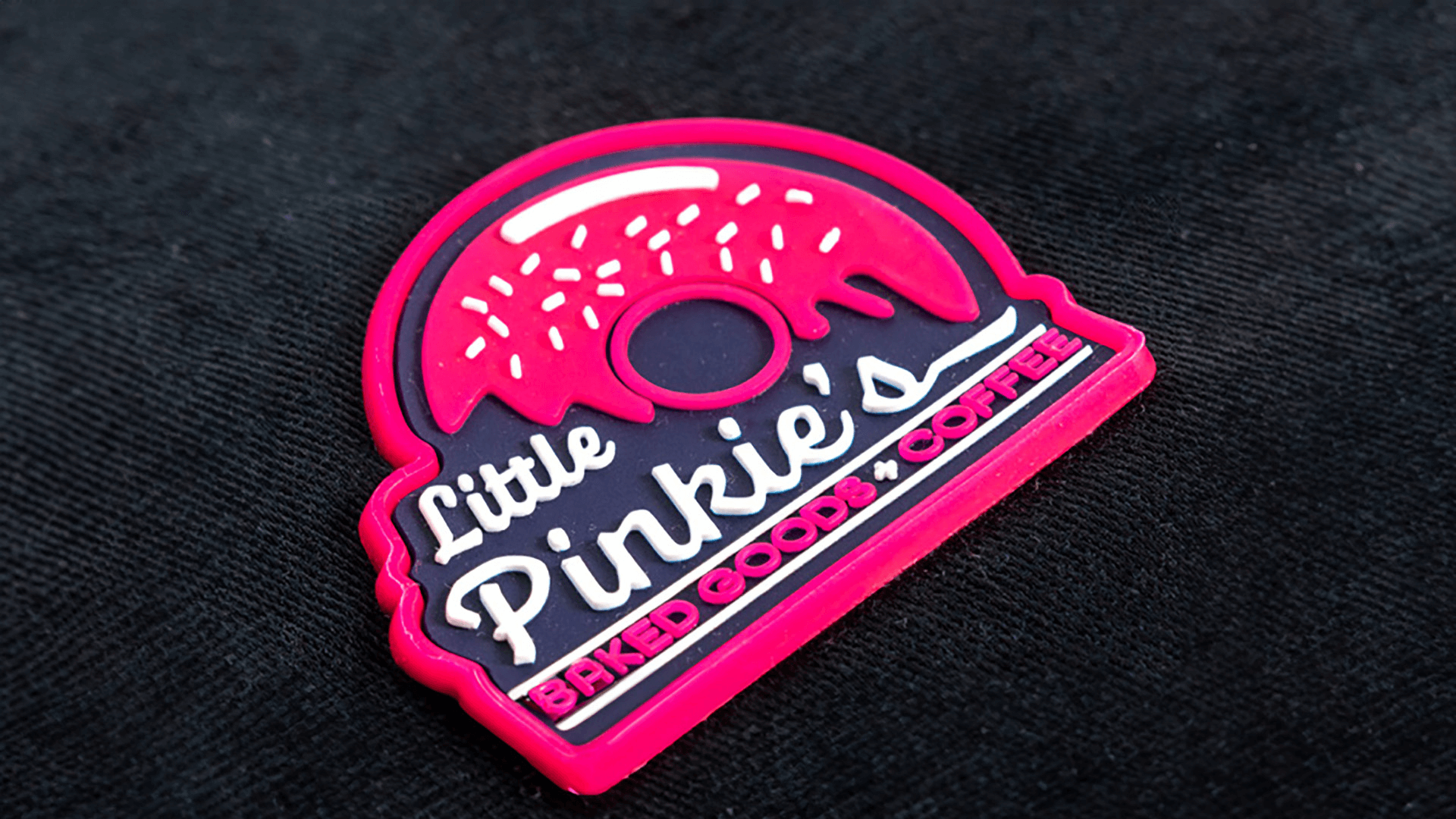 A vibrant PVC patch with a donut design, featuring "Little Pinkie's Baked Goods & Coffee" in white and pink lettering, with a playful donut shape on a black background