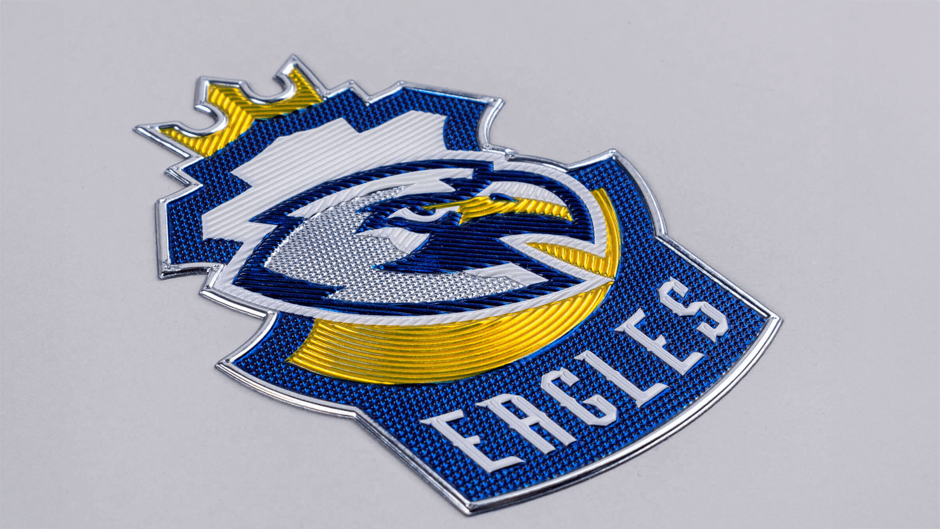 A detailed eagle emblem with a blue and yellow design, featuring the word "EAGLES" at the bottom and a crown-like shape at the top, against a gray background