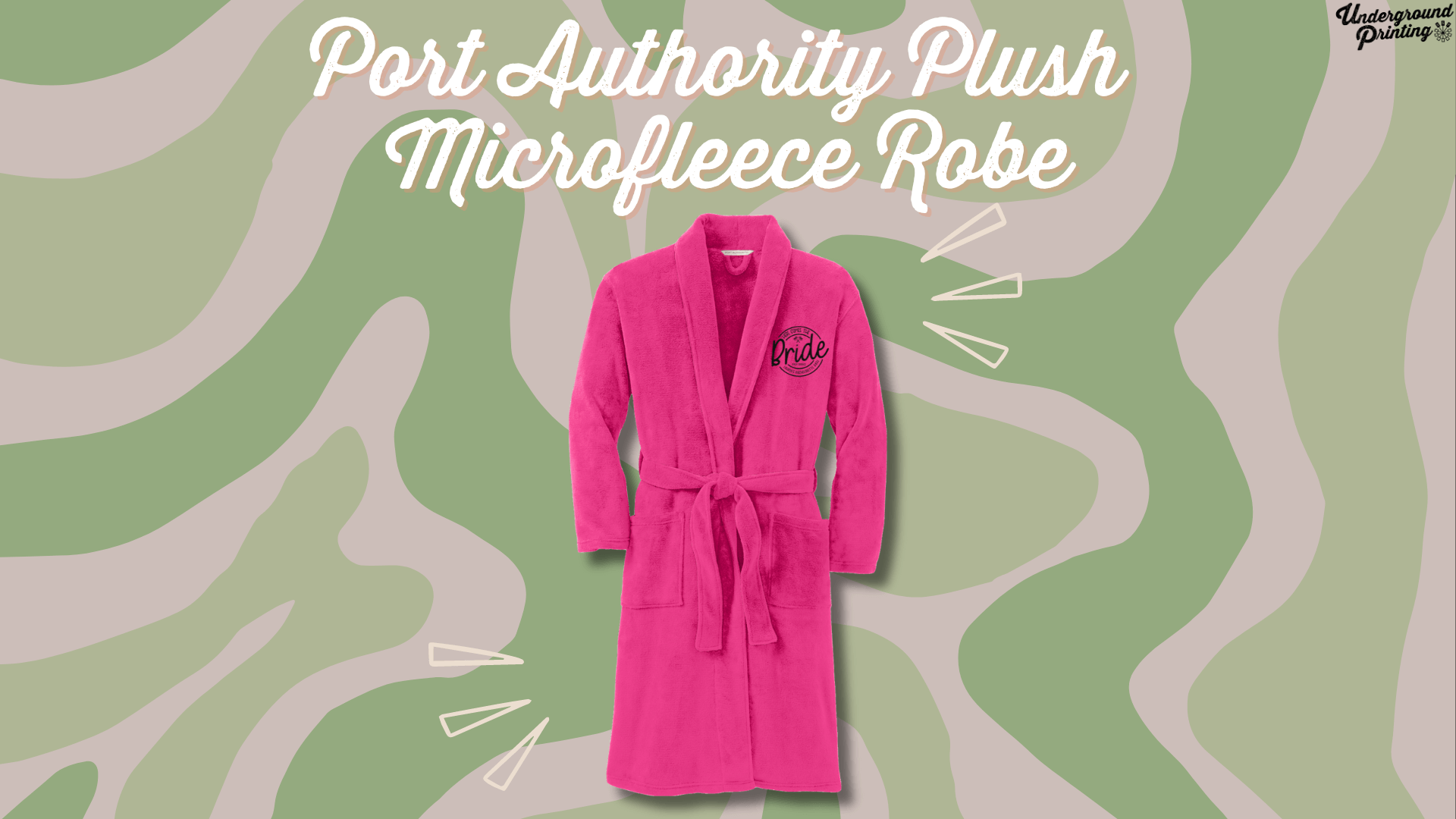 A bright pink, soft microfleece robe with a "Bride" emblem on the chest. The robe is set against a green and beige camouflage-style background, with decorative accent lines highlighting the product.