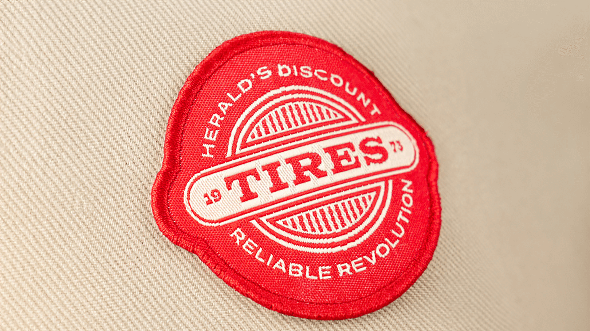 A bright red, circular woven patch with a tire-themed logo for "Herald's Discount Tires," featuring the text "Reliable Revolution" along the edge. The patch is stitched onto a beige fabric background