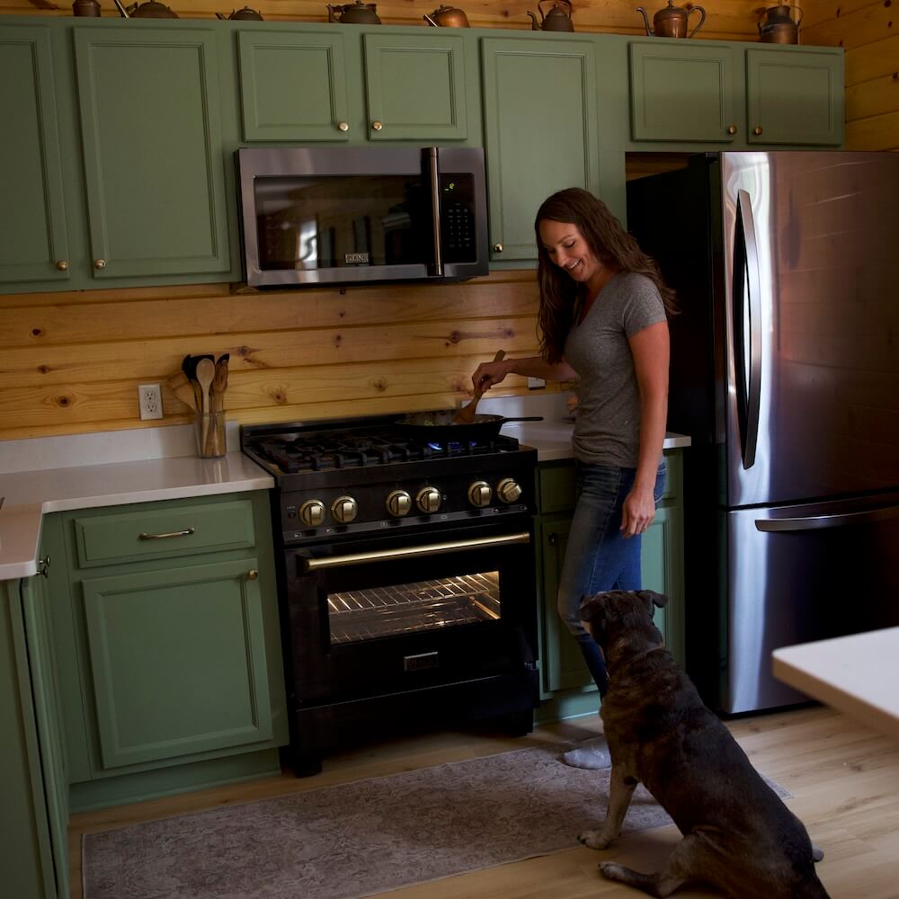 A woman cooking food on a range below a microwave range hood while a dog watches