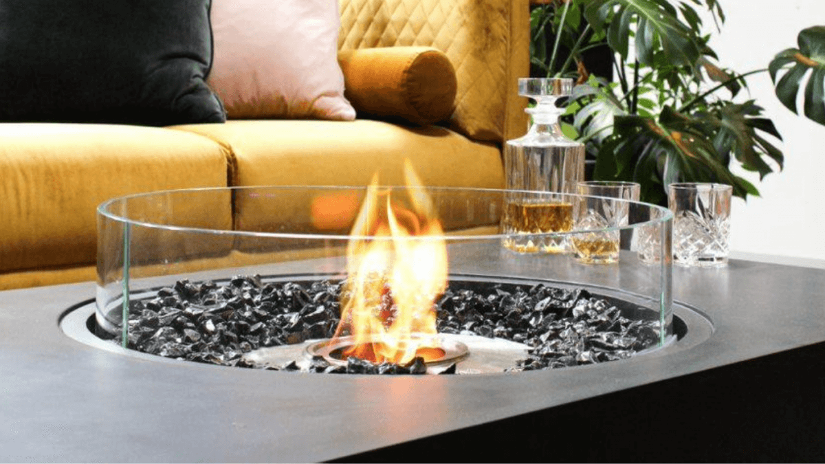 a round fire pit table with flames with a wind guard with a couch and plants in the back