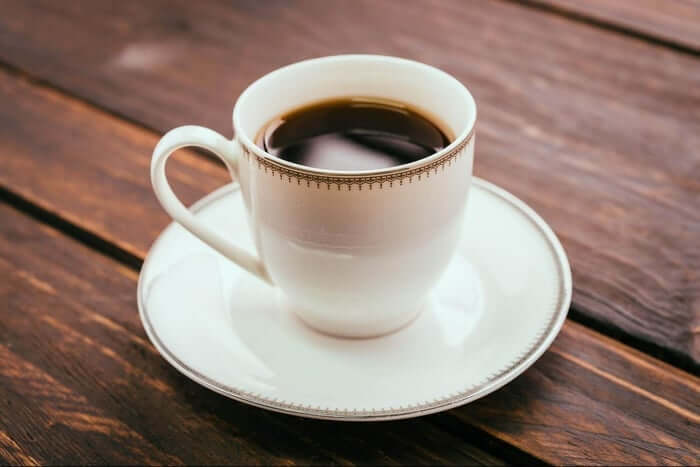 White porcelain cup filled with black coffee on saucer over wooden table.