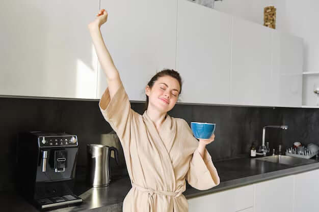 A young woman in a bathrobe, stretching joyfully while holding a morning coffee