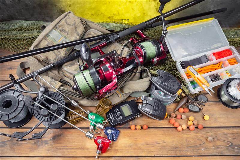 Assortment of fishing tackle, reels, bite alarms, and bait organized on a wooden surface.