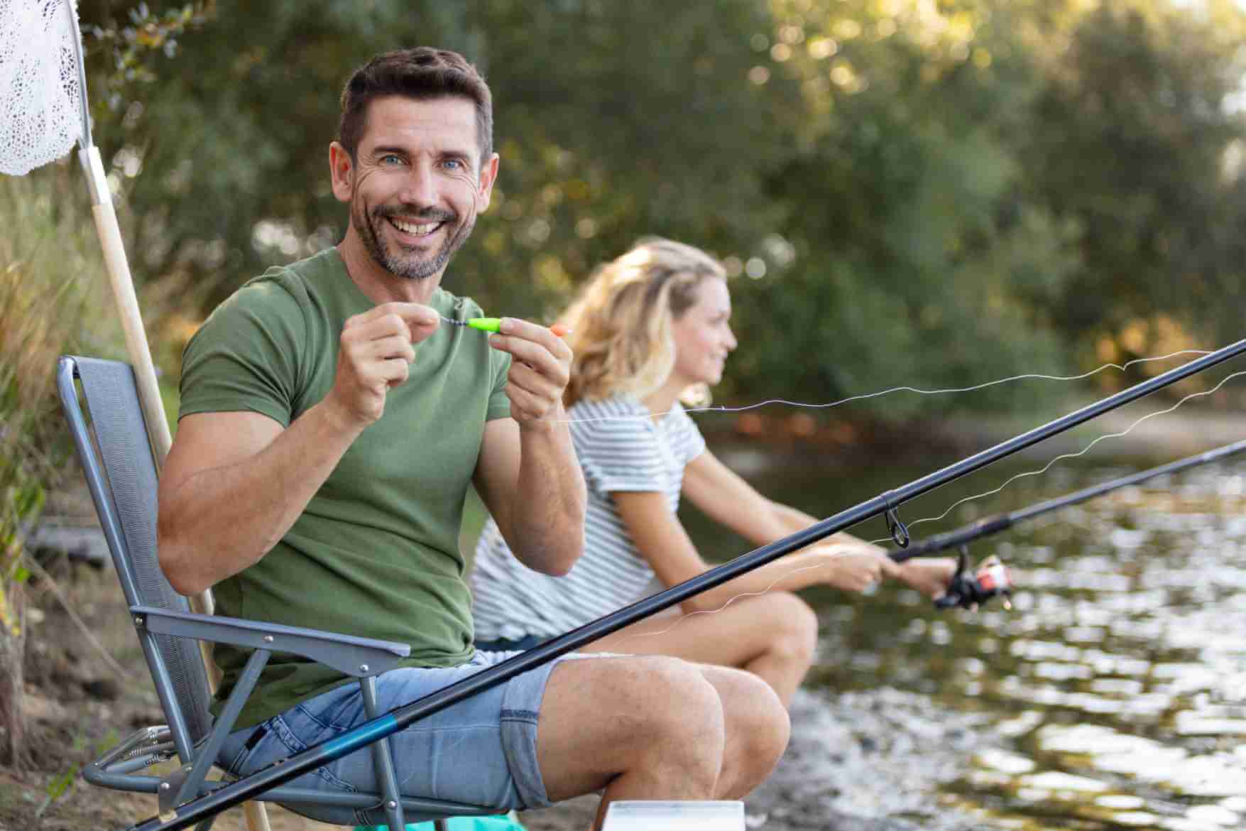 Man smiling while baiting a hook beside a river, woman fishing in background.