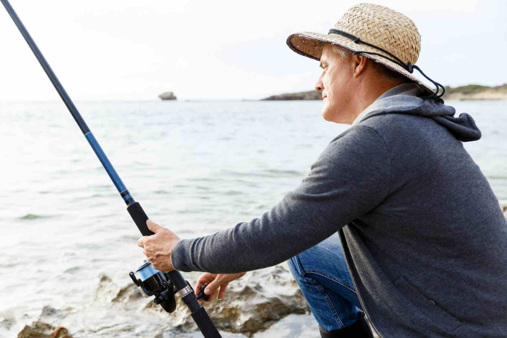 Man in a straw hat fishing from a rocky shoreline, holding a rod and looking at the water.