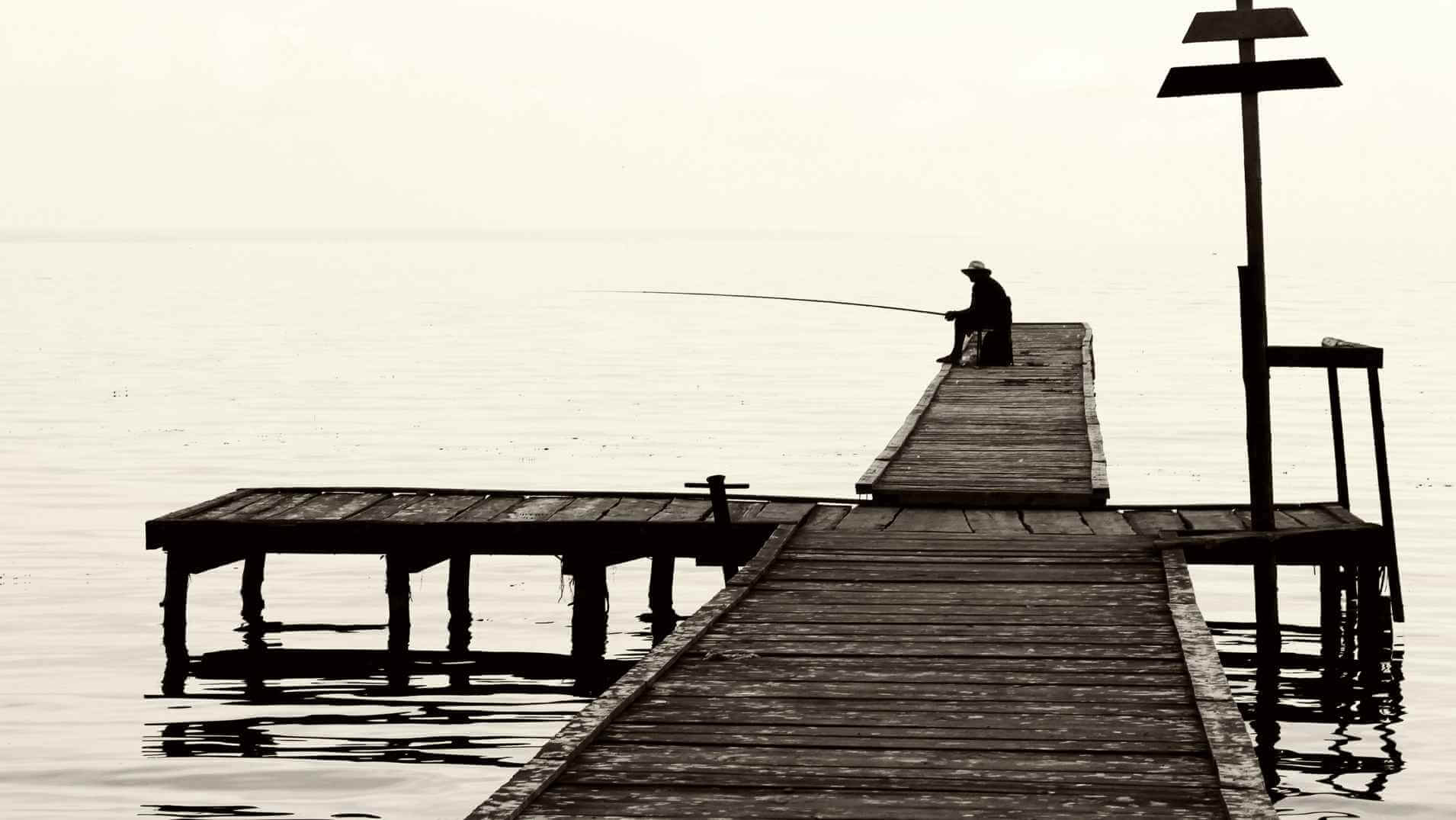 Angler fishing at the end of a wooden pier, casting a rod over calm seawater on a bright clear day.