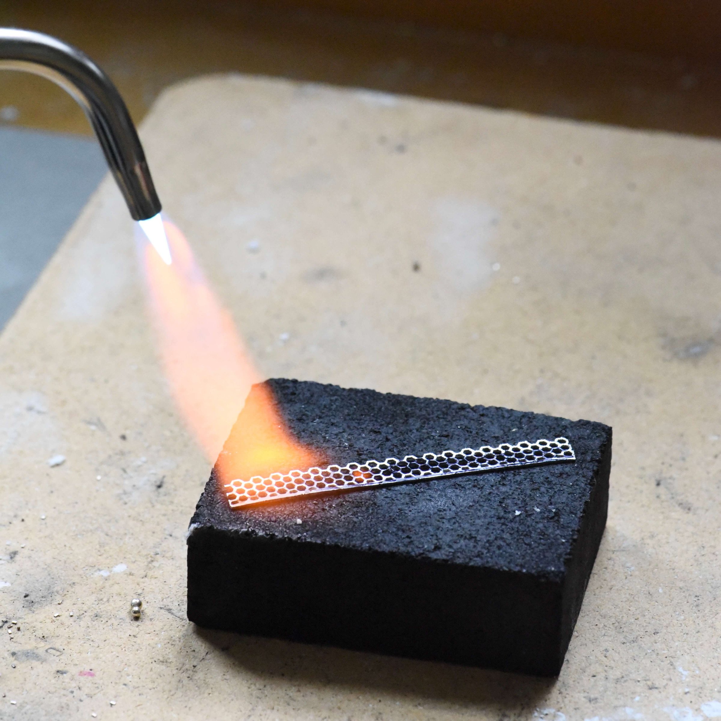A ceramic soldering board with a charcoal block on top, as a jeweler anneals a piece of silver using a torch flame.