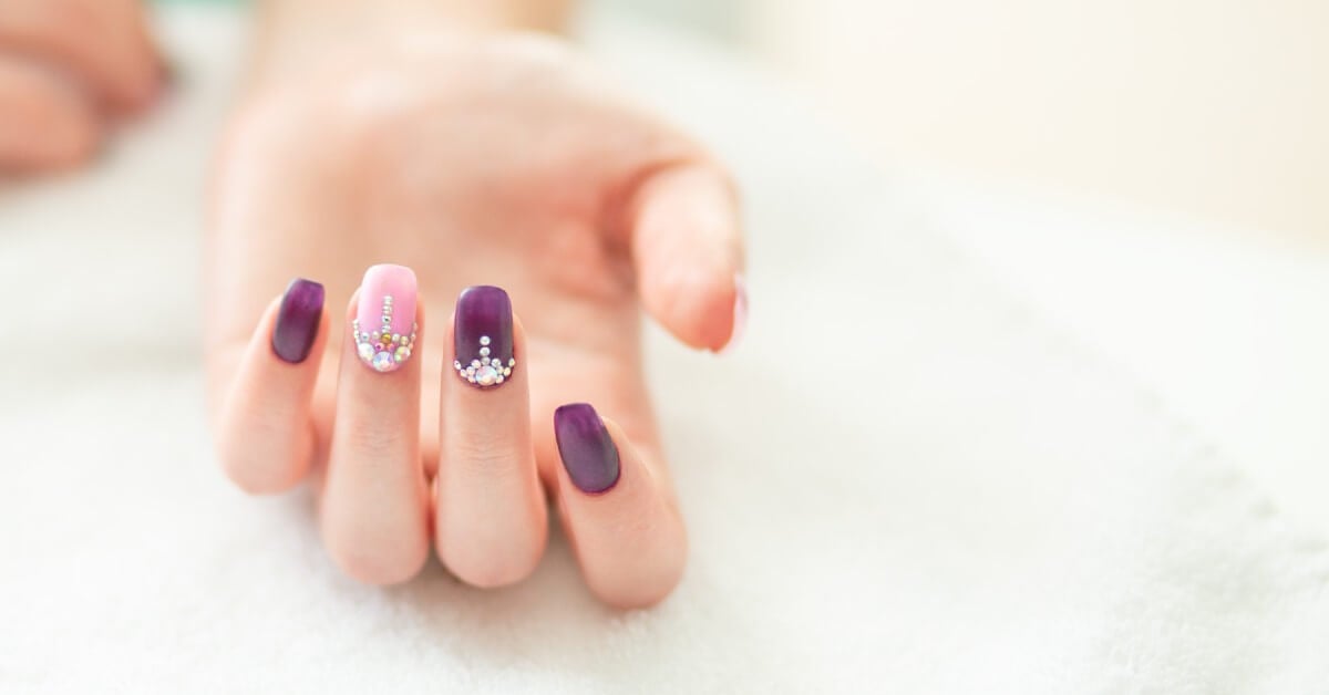 A hand with three nails painted plum-purple while the ring fingernail is pale pink; the middle two nails have rhinestones.