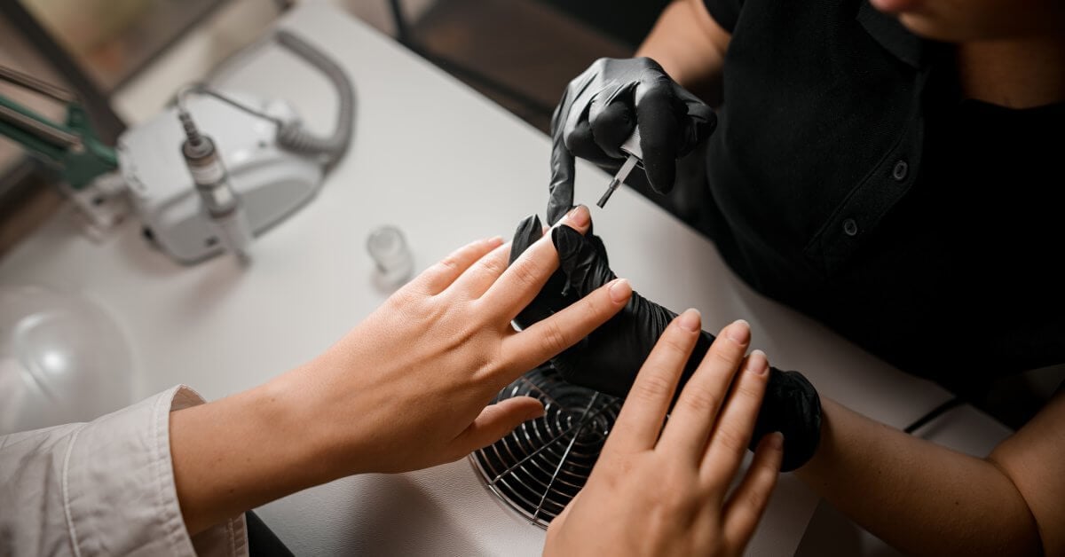 An overhead look of a manicurist holding someone’s finger steady as they paint a clear coat of polish on the nail.