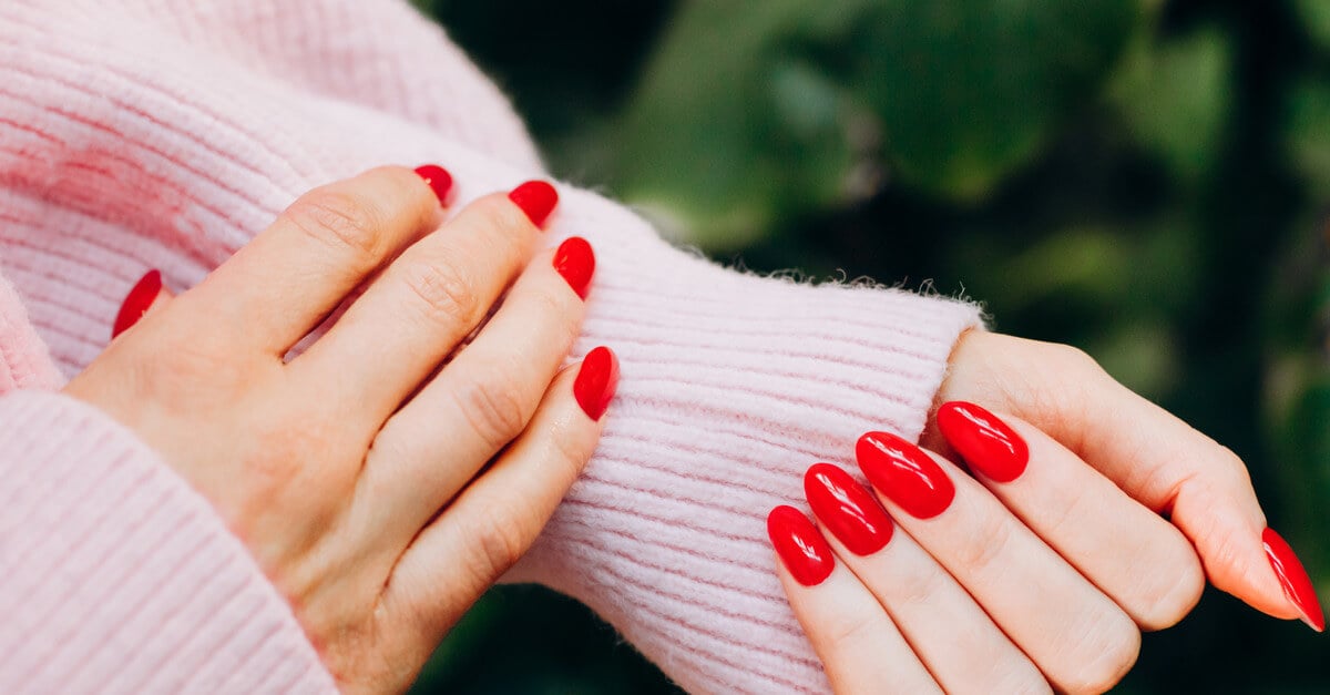 A close-up of female hands with cherry red almond nails and a woolen pink sweater on a green background.