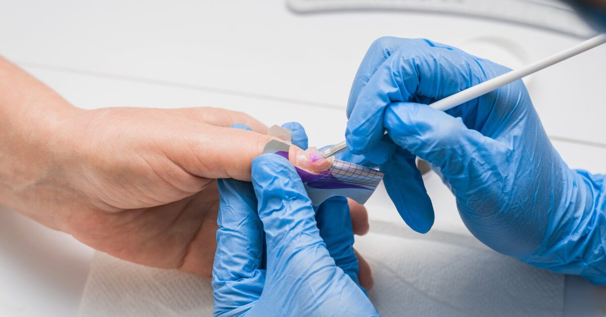 A pair of hands in blue rubber gloves hold a nail art brush and another hand steady to apply product to the nail.