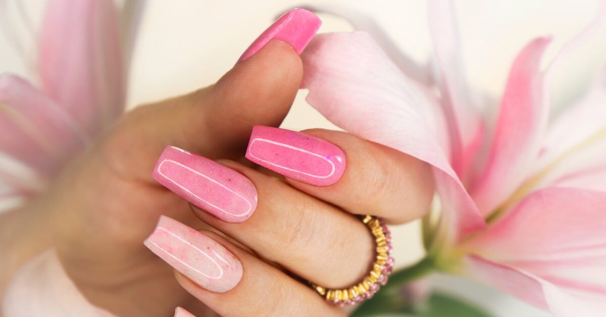 A close-up of someone's hands with long pink and fine glitter nails in a square shape. They're wearing a pink diamond ring.