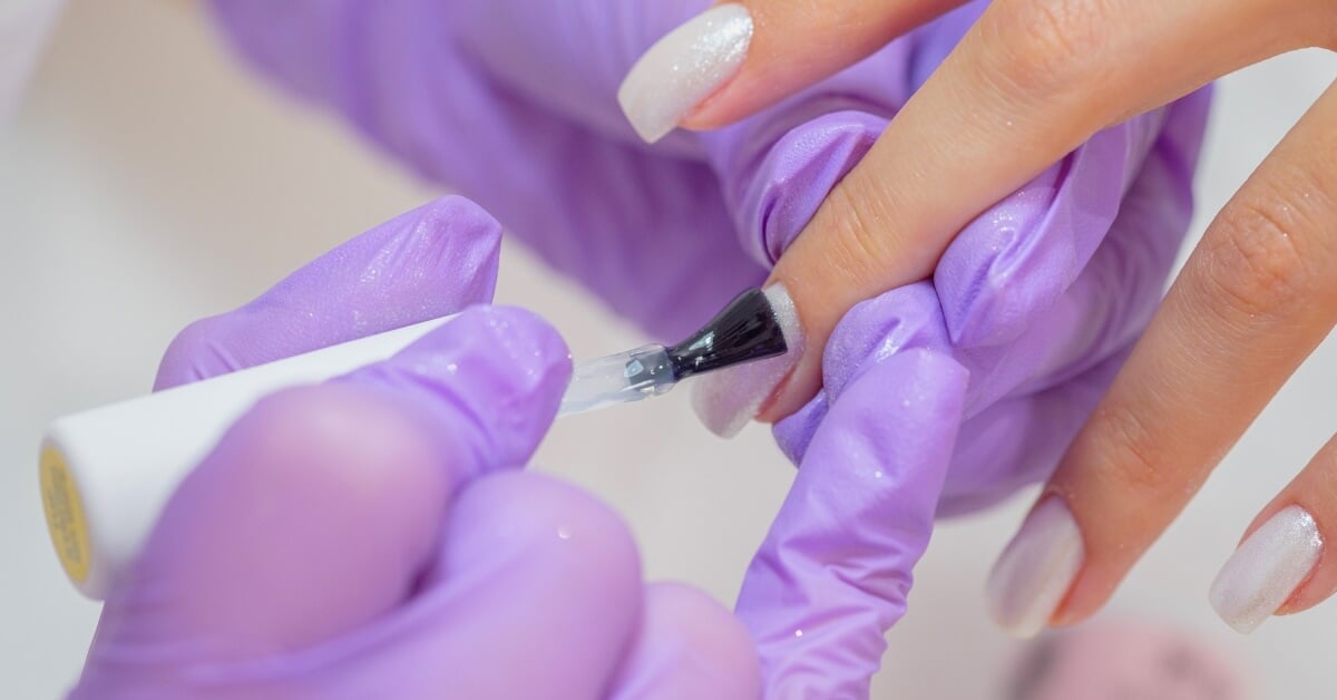 A pair of hands wearing purple rubber gloves holds a manicured hand and polish brush to paint the nail.