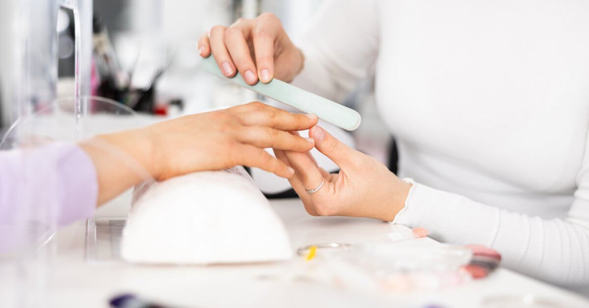 A manicurist shapes a young person's nails with a white nail file during a nail salon manicure session.