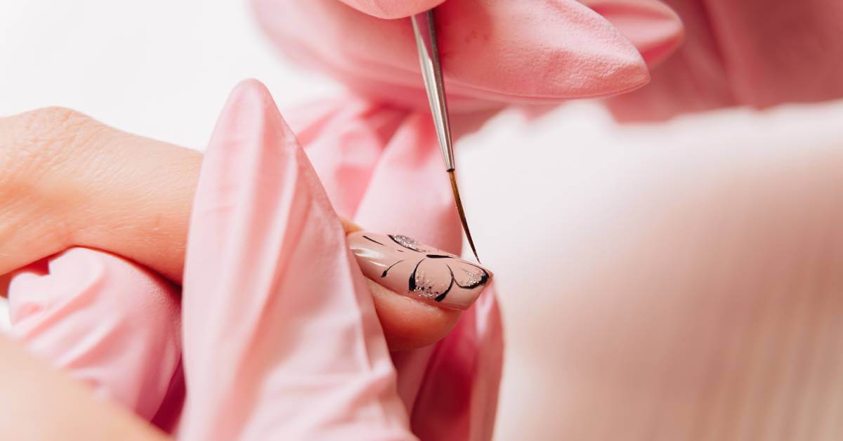 A nail tech’s gloved hand holds a client’s finger steady and uses a nail art brush to create an intricate design on the nail.