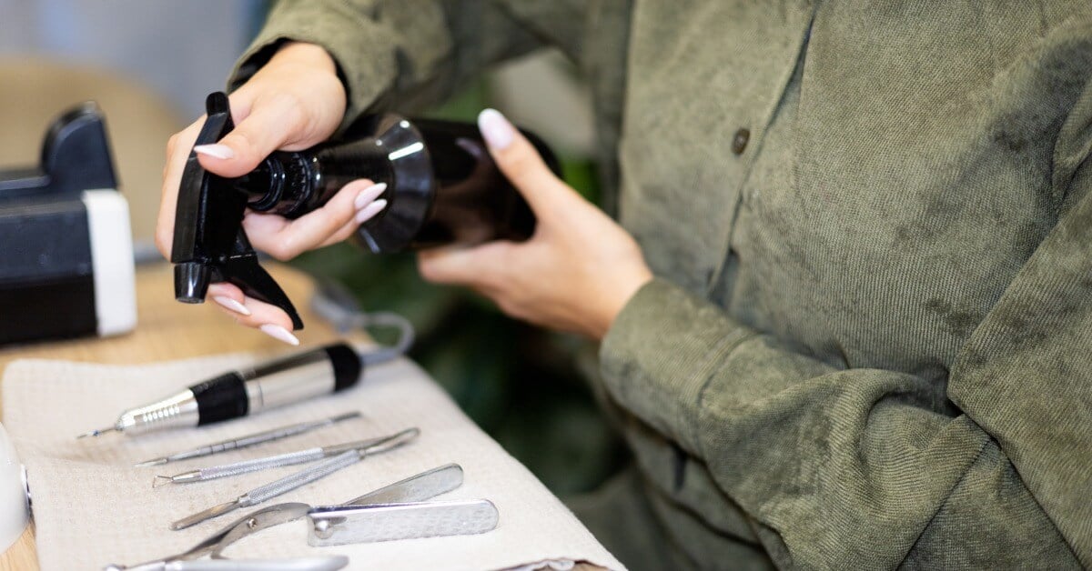 Two hands hold a black spray bottle over various manicure tools, including nail cutters and cuticle pushers, on a desk.