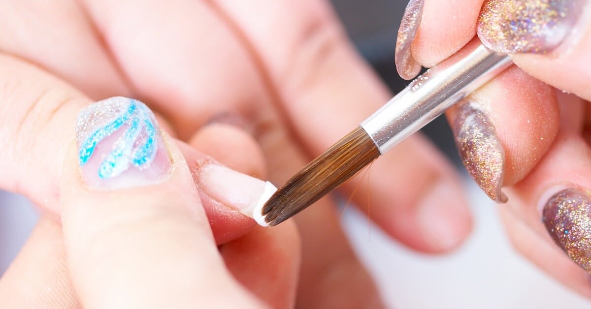 A manicurist wearing various nail polish designs corrects acrylic tips by using a small brush with acrylic on the end.