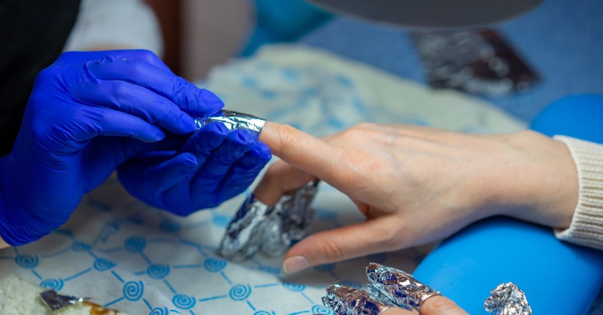 A technician wraps foil around a customer's fingernails to remove polish from their hands during a manicure.