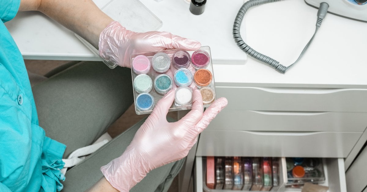 A manicurist wearing gloves sits at a white desk while organizing their manicure nail equipment inside a cabinet.
