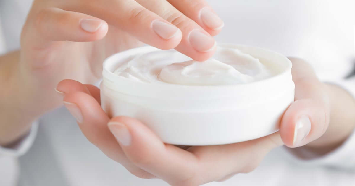 A person in a white long-sleeve shirt applies moisturizing cream from a small white round jar to their hands.