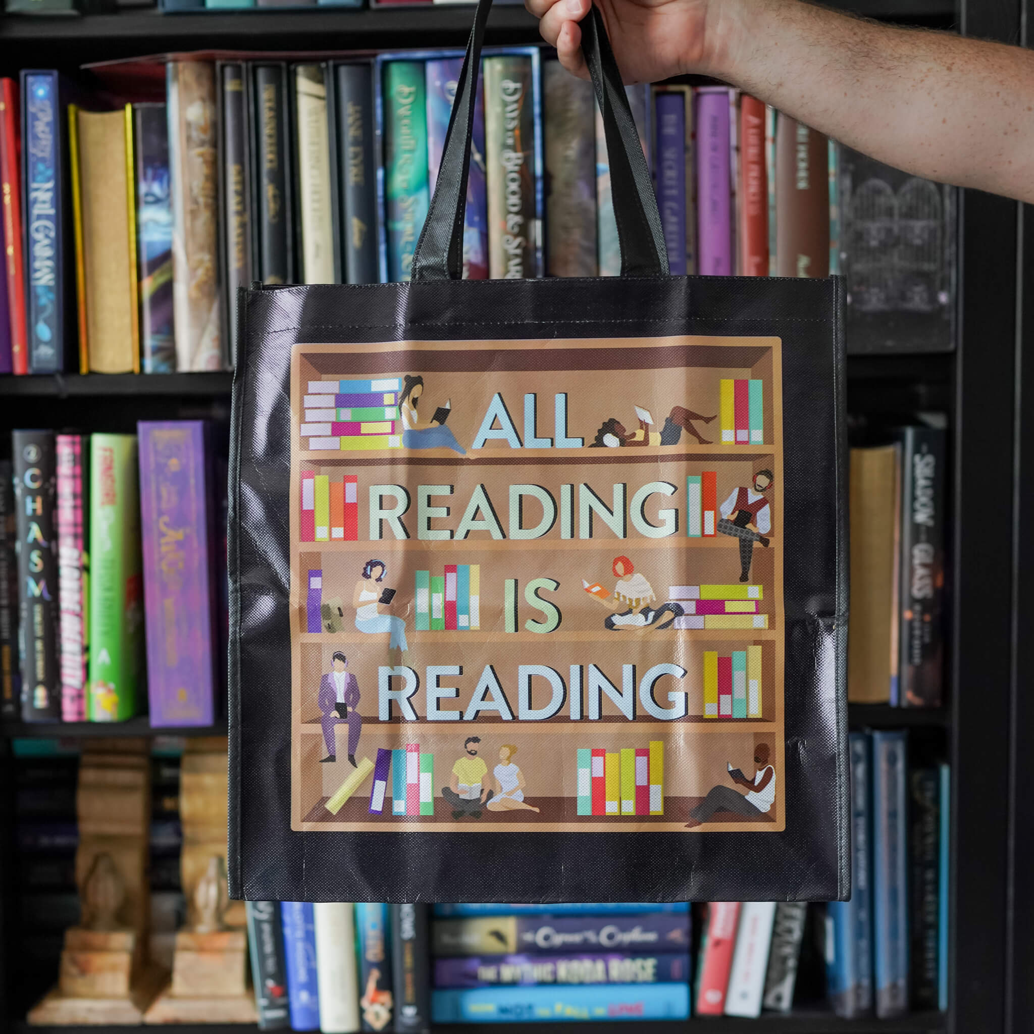 Black reusable grocery bag that says "All reading is reading" on a book shelf with people sitting on it reading/listening 