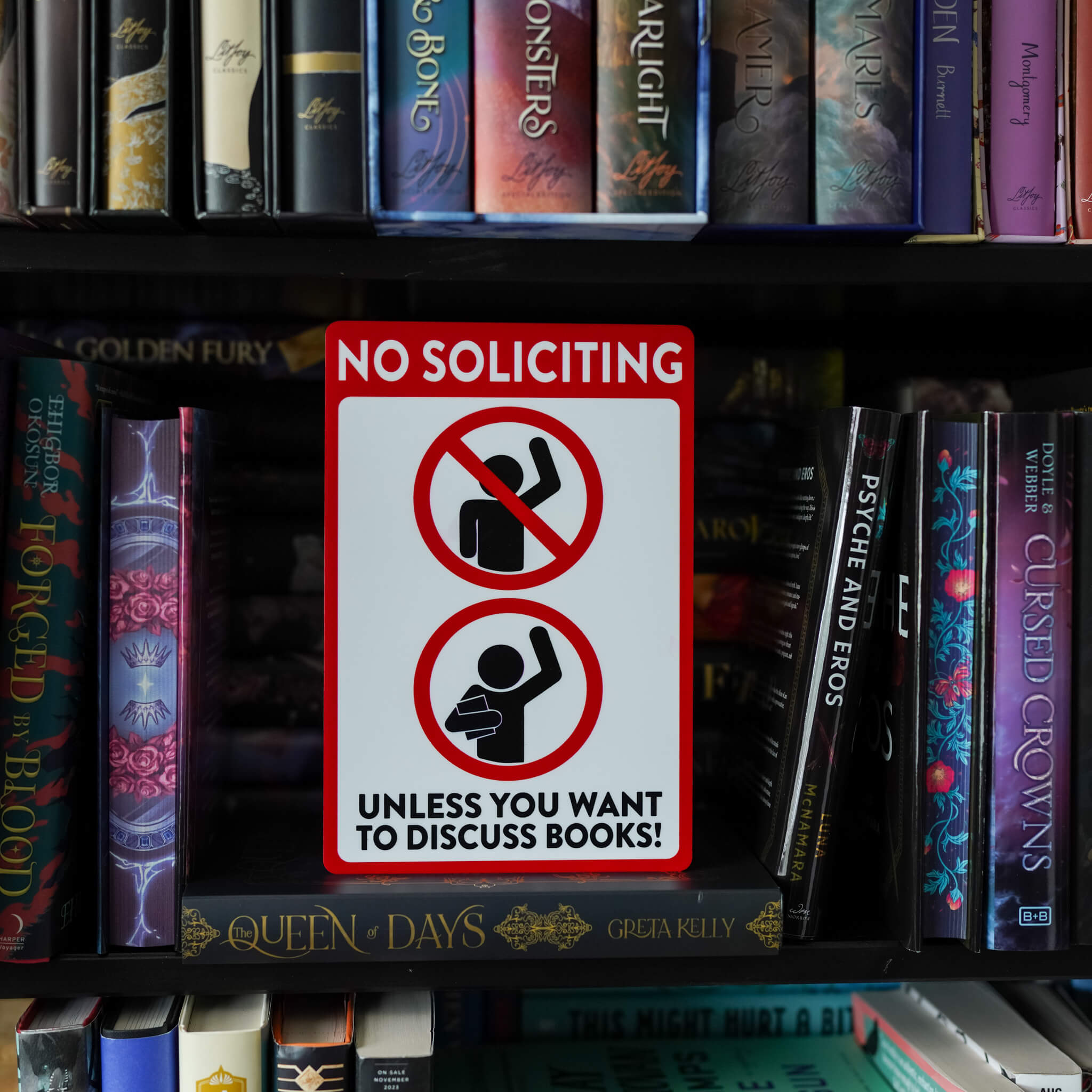 White sign with red border and symbols, black stick figures, reading "No soliciting unless you want to discuss books"