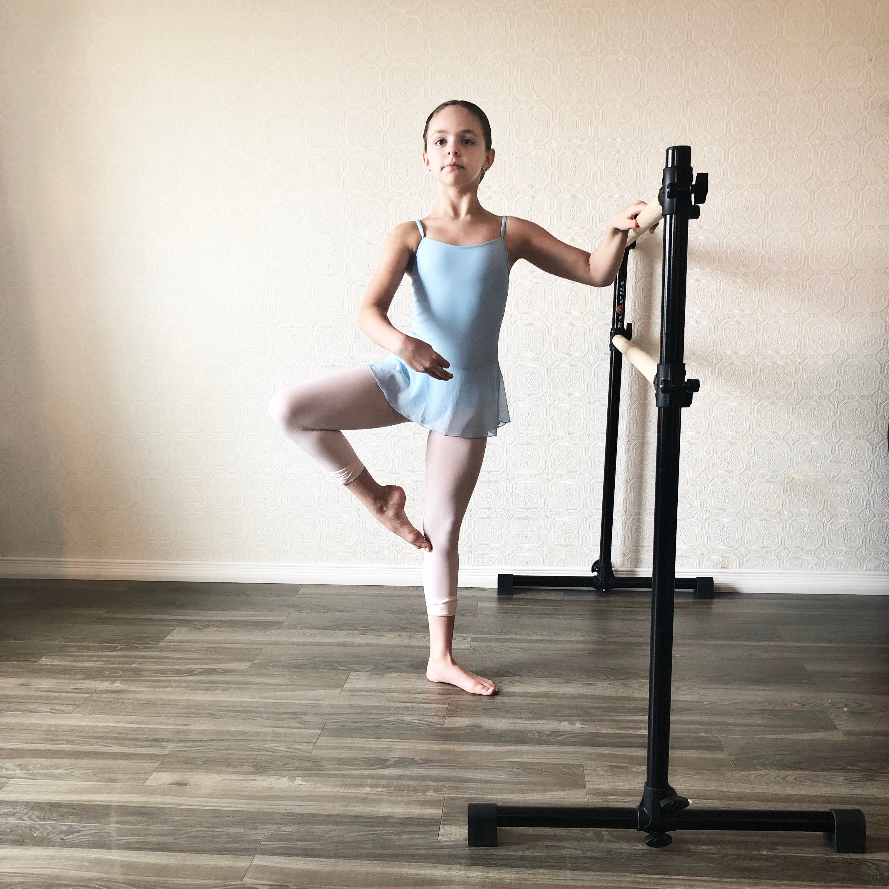 Young girl in ballet attire practicing ballet pose at a black free standing ballet barre for children in home studio