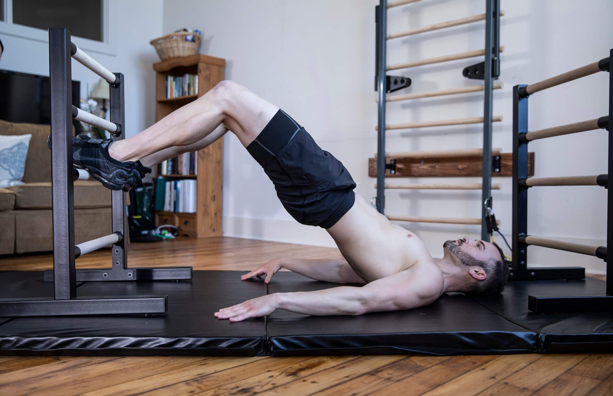 A man performs a glute bridge exercise at home using professional ballet bars for home equipment from Vita Barre. He rests his feet on the lower rungs of a black freestanding barre while lying on a padded mat in a bright studio-style living space. The setup showcases durable, USA-made equipment for ballet designed for strength, balance, and core training.