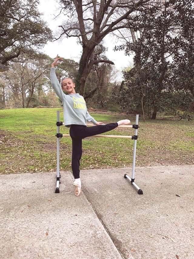 A young dancer practices barre ballet outdoors using a portable ballet barre, smiling while stretching her leg and posing gracefully in a peaceful park setting.