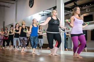 Group of women participating in a barre workout class using a ballet barre and light hand weights. They are performing synchronized movements in a bright studio with mirrors and wooden floors, demonstrating strength, balance, and coordination during a winter fitness session.