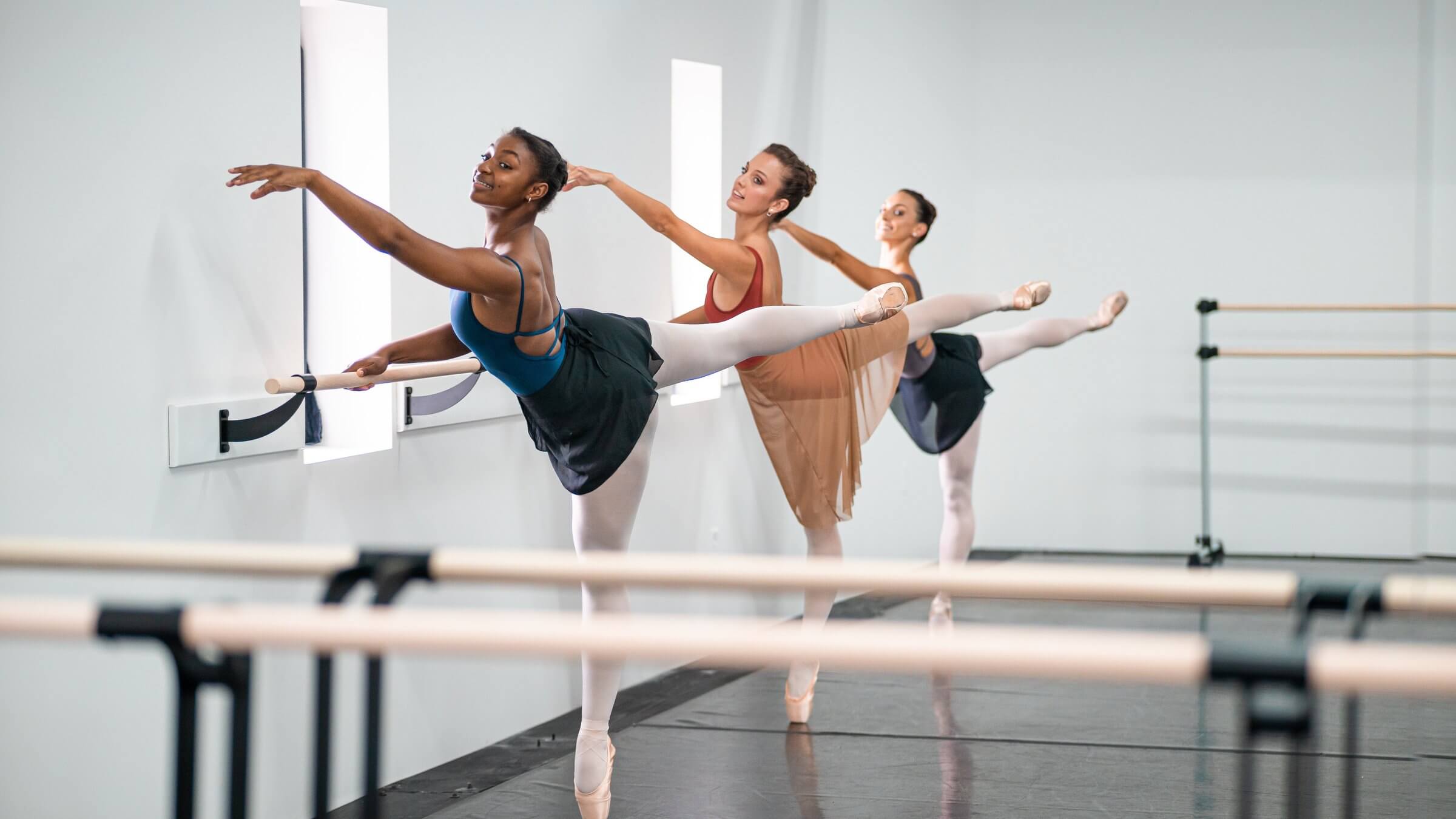 Three ballet dancers performing arabesque at dance barre in modern studio with adjustable ballet barres