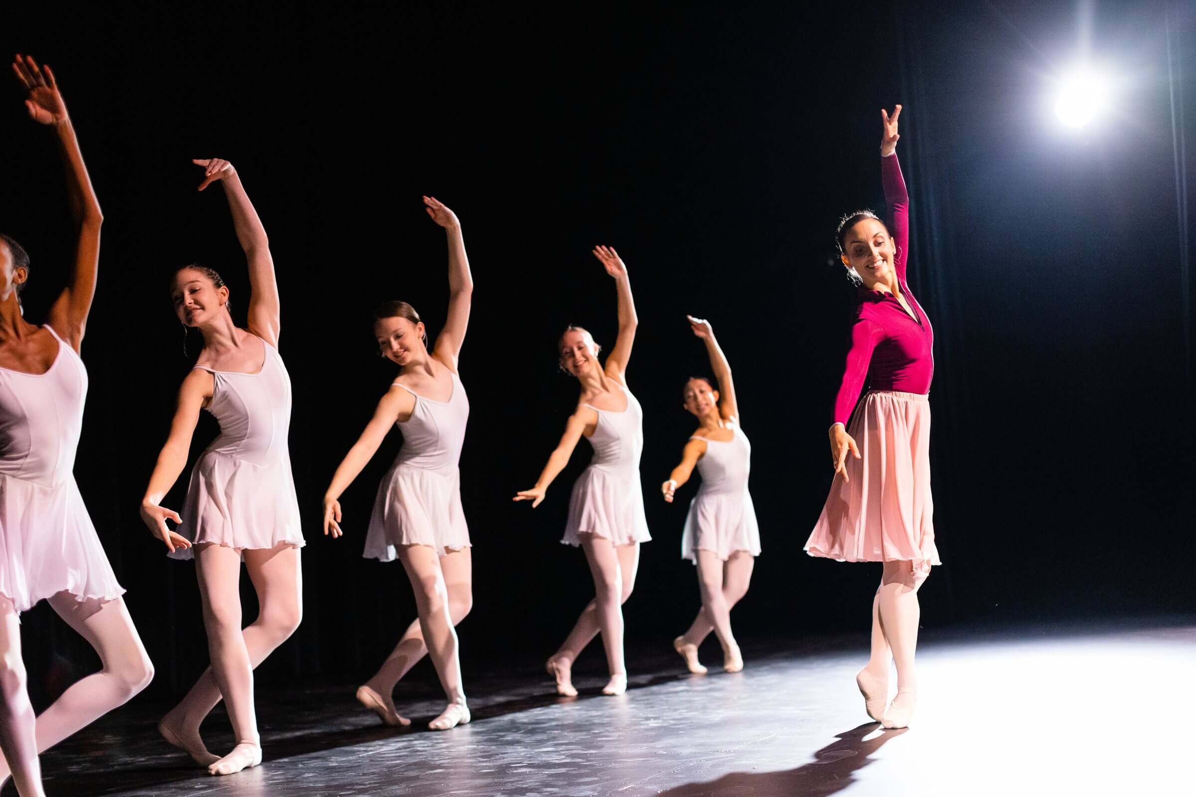 Group of six female dancers in pink and white leotards with flowing skirts performing synchronized dance barre movements on stage under dramatic spotlight, demonstrating proper form and technique during ballet-inspired fitness routine
