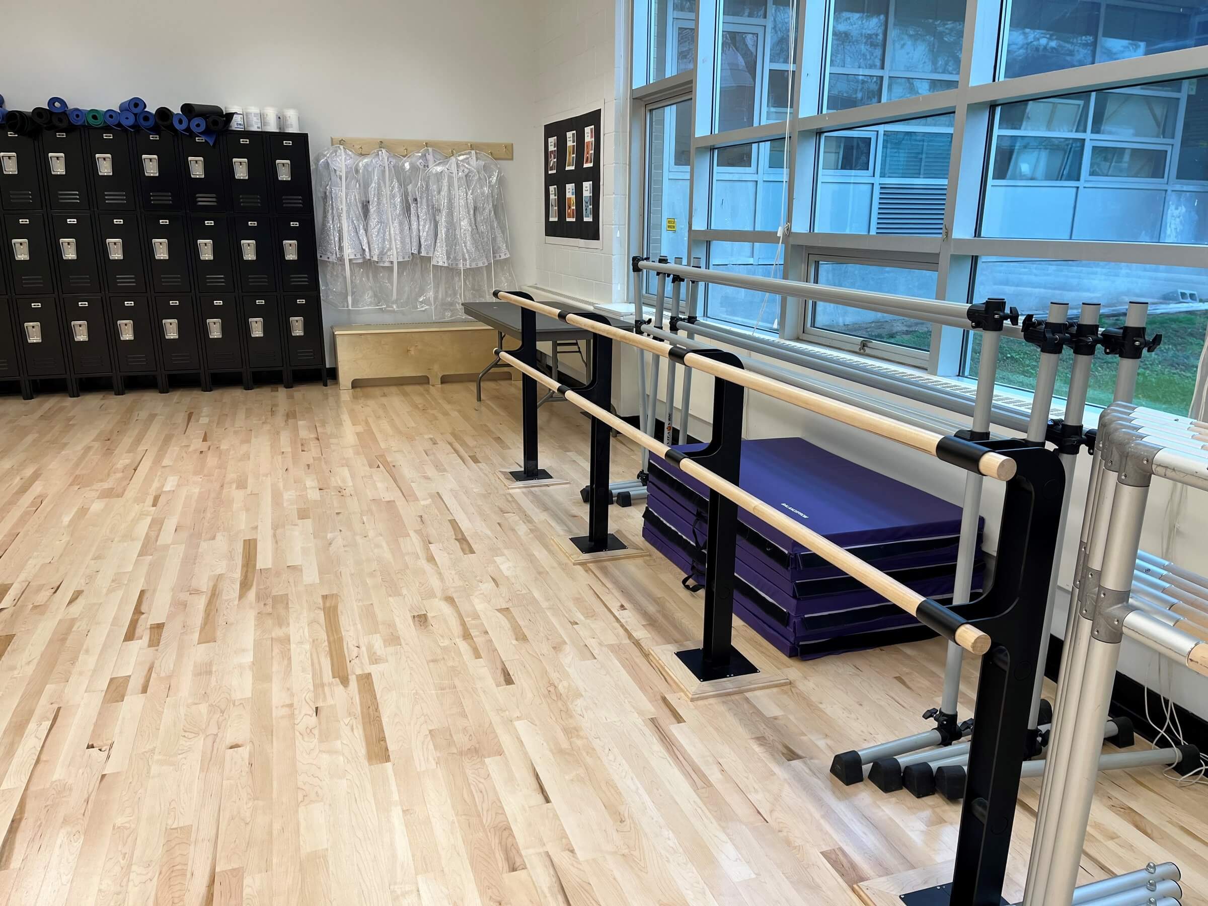 A bright studio with wooden floors, black lockers, and several freestanding ballet barres positioned near large windows. Folded purple mats are stacked beneath the windows, and clear garment bags hang on hooks along the back wall. The space demonstrates how professional equipment like these ballet barres can also inspire a functional barre for home setup.