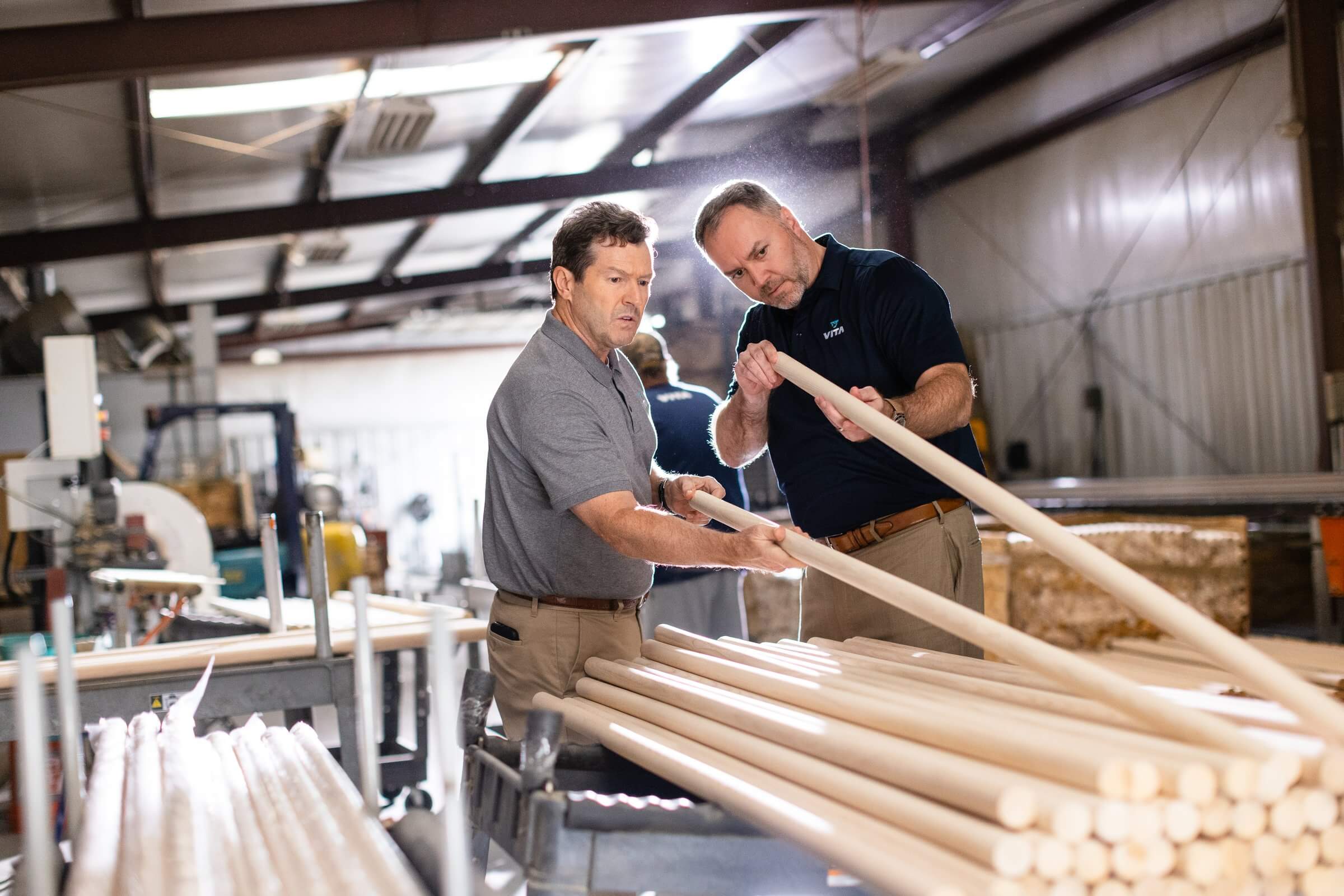 Two men inspect wooden ballet barre poles inside a workshop, carefully checking the finish and quality of the materials used for professional studio barres