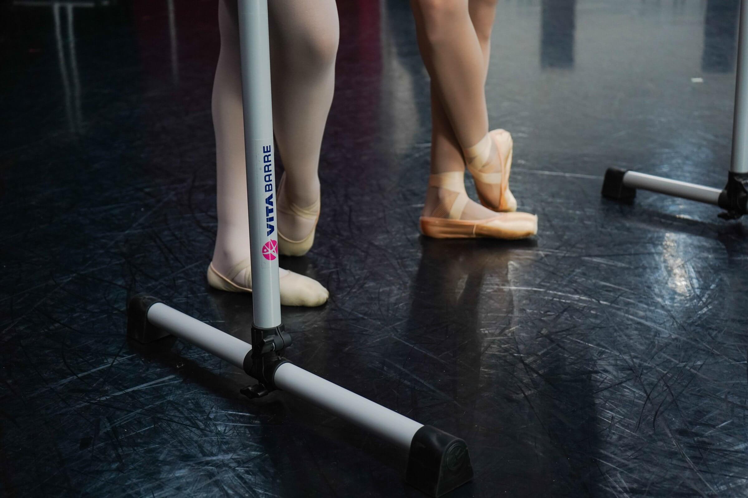 Close-up of two ballet dancers practicing on a Vita Barre portable barre, showing their pointe shoes and elegant posture — high-quality equipment for ballet designed for strength, balance, and precision training.