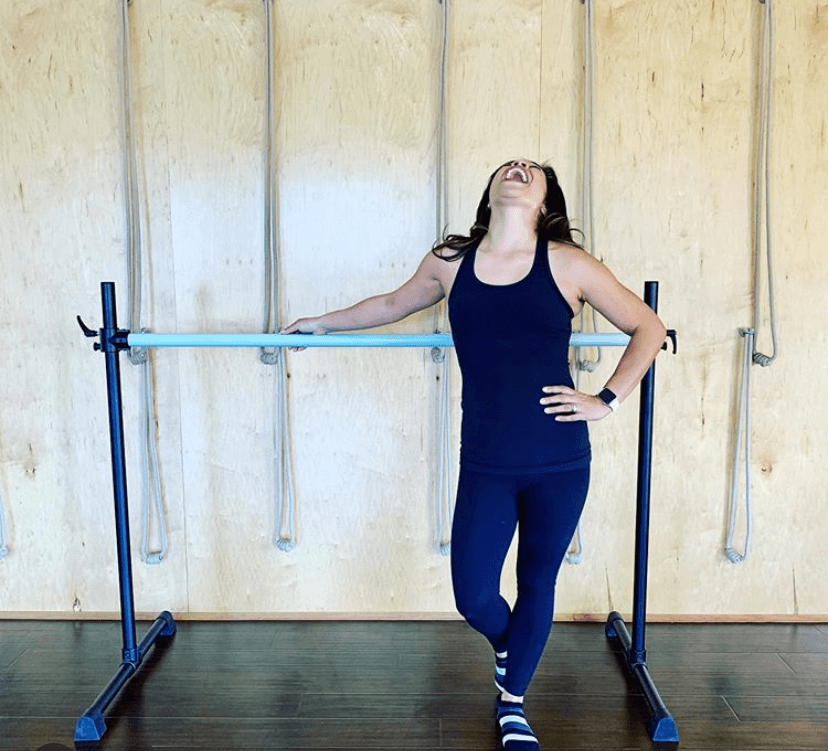 A woman stands at a professional ballet barre in a bright studio, smiling and relaxed after a barre ballet workout, showcasing strength, confidence, and balance.