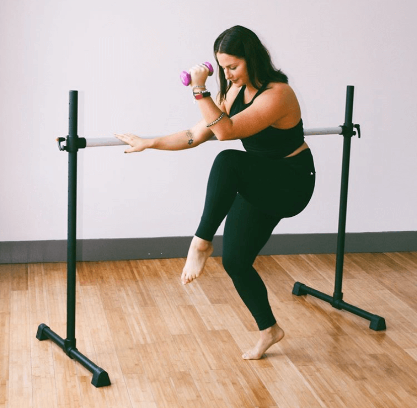 A woman performs a barre fitness exercise using a professional ballet barre, holding a small dumbbell while lifting one leg in balance. She’s barefoot on a wood studio floor, demonstrating strength and stability during a barre ballet workout.