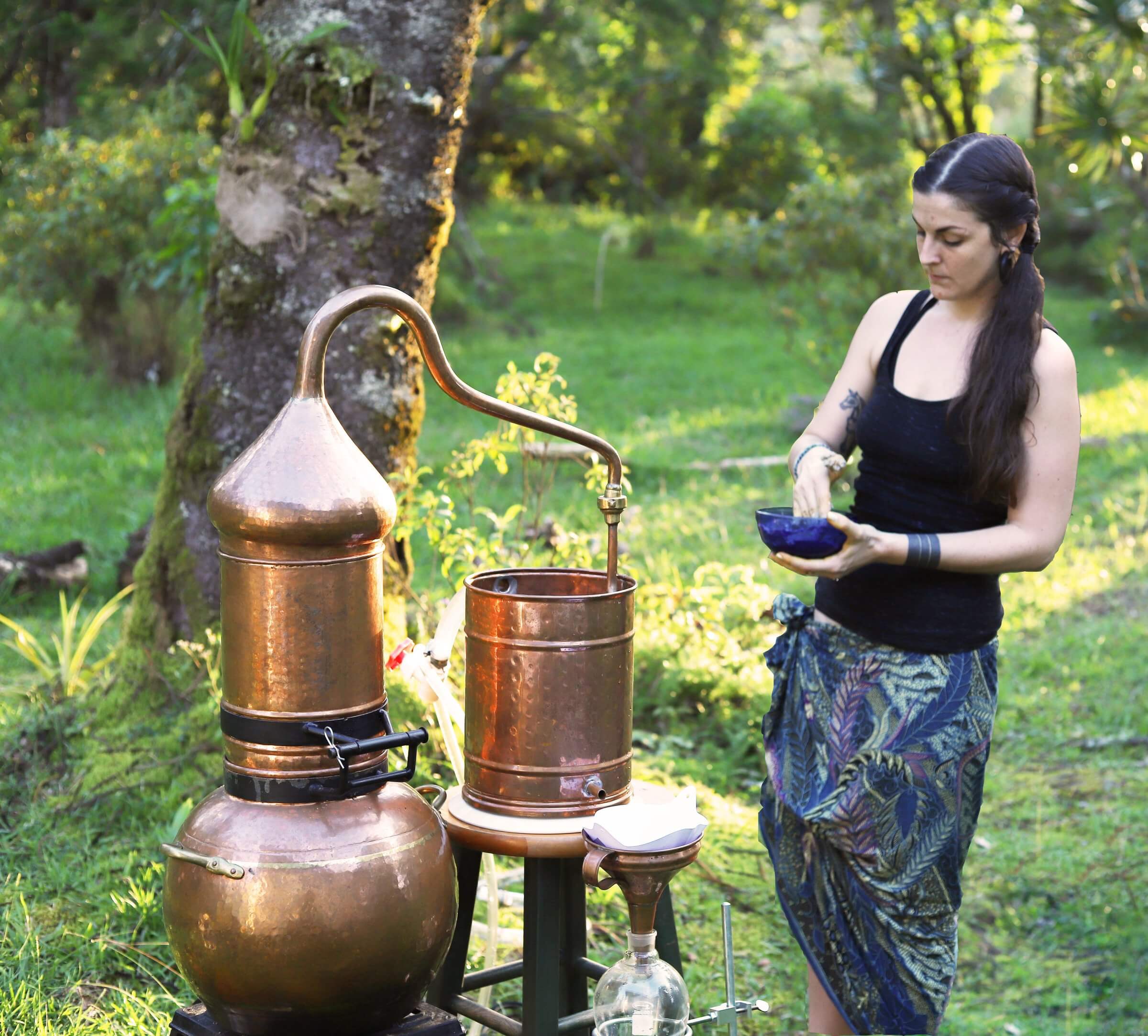 A woman standing next to a copper alembic still in a garden, holding a bowl of rye flour paste to seal the still