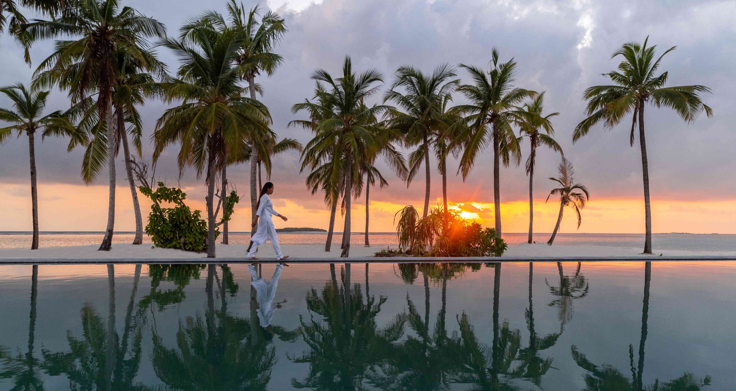 A view of a woman past the infinity pool at Alila Kothaifaru Maldives at sunset, framed by the ocean and palm trees. 